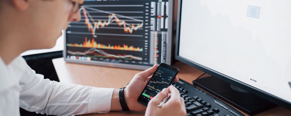 Over the shoulder view of and stock broker trading online while accepting orders by phone. Multiple computer screens ful of charts and data analyses in background